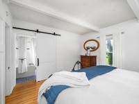 Bright Orono bedroom at 405 S Old Crystal Bay Road with white walls, a bed with a white and blue blanket, wooden floor, dresser with mirror, window with curtains, and a sliding barn door leading to the bathroom. - Beise Real Estate