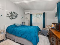 A bedroom on Rainbow Avenue in Maple Plain features a bed with a teal comforter, nightstand, dresser, armchair, wall decor, a window with teal curtains, and an air conditioner above the window. - Beise Real Estate