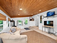 A cozy living room at 405 S Old Crystal Bay Road in Orono features a wood-paneled ceiling, white walls, a beige sofa, TV on a stand, large windows, and a door to the outdoors. Mounted fish decor sits above the TV. - Beise Real Estate