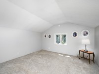 A small, carpeted room with sloped ceilings, a window overlooking Old Crystal Bay Road in Orono, a table with a lamp, and four framed photos hanging on the wall above the window. - Beise Real Estate