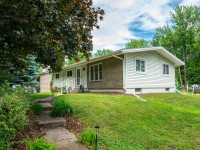 Single-story house with white siding, front porch, and large windows on Rainbow Avenue in Maple Plain, surrounded by a green lawn, trees, and landscaped shrubs on a partly cloudy day. - Beise Real Estate