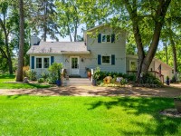 Charming two-story white house with blue shutters on 405 S Old Crystal Bay Road, featuring a front porch, outdoor seating, and a large green lawn surrounded by trees in sunny Orono. - Beise Real Estate