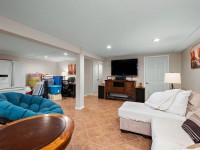 A basement room on Rainbow Avenue in Maple Plain features tile flooring, a sectional sofa, wall-mounted TV, small desk area, refrigerator, storage bins, and neutral walls with recessed lighting. - Beise Real Estate
