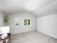 A small, carpeted bedroom with gray walls at 405 S Old Crystal Bay Road, features a window overlooking greenery, a double-door closet, a lamp on a wooden table, and a painting and wreath on the wall. - Beise Real Estate