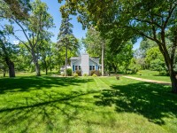 A small white house with a chimney sits in the center of a large, well-maintained green lawn on Old Crystal Bay Road in Orono, surrounded by mature trees under a clear blue sky. - Beise Real Estate