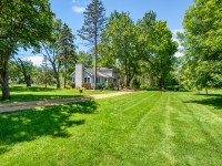 A house with light siding sits on a large, well-manicured lawn along Old Crystal Bay Road in Orono, surrounded by tall trees under a sunny, blue sky. - Beise Real Estate