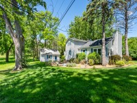 A light gray house with a detached garage sits on a grassy lot surrounded by trees under a clear blue sky at 405 S Old Crystal Bay Road in Orono. - Beise Real Estate