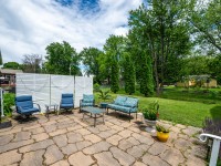 Stone patio on Rainbow Avenue in Maple Plain with blue cushioned chairs, a glass table, potted plants, and a white privacy screen, overlooking a grassy yard framed by trees and neighboring houses. - Beise Real Estate