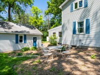 A small backyard on Old Crystal Bay Road features a round metal table with four chairs on mulch, beside a light gray house and detached garage with blue shutters and door, all surrounded by trees and greenery. - Beise Real Estate