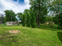 A grassy backyard on Rainbow Avenue in Maple Plain features a circular fire pit, several tall trees, and a chain-link fence separating the yard from neighboring houses. - Beise Real Estate