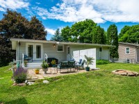 Single-story house on Rainbow Avenue in Maple Plain with a back patio featuring outdoor furniture, potted plants, and a fire pit, surrounded by grass and trees under a partly cloudy sky. - Beise Real Estate