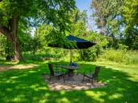 A round table with four chairs and a large black umbrella sits on green grass along Old Crystal Bay Road in Orono, surrounded by trees and lush foliage in a sunny outdoor setting. - Beise Real Estate