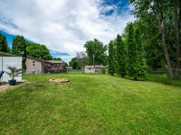 A grassy backyard on Rainbow Avenue with a fire pit, patio area with chairs, trees along the fence, and houses in the background under a partly cloudy Maple Plain sky. - Beise Real Estate