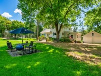A white house with a front porch and detached garage sits on a large grassy yard along Old Crystal Bay Road in Orono, featuring outdoor seating, a grill, and an umbrella under tall green trees on a sunny day. - Beise Real Estate