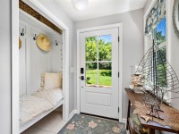 A small entryway at 405 S Old Crystal Bay Road features a built-in bench with pillows, wall hooks holding a hat, a glass door opening to a green yard, and a table with a decorative wire sailboat and shells. - Beise Real Estate