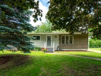 Single-story house on Rainbow Avenue in Maple Plain, featuring a small front porch, light siding, brick accent, and large windows—surrounded by green lawn, trees, and beautiful landscaping. - Beise Real Estate