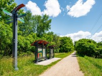 A gravel trail runs beside a shaded rest area with info boards and a signpost along the Luce Line Trail, surrounded by green trees under a partly cloudy sky. - Beise Real Estate