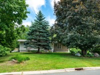 Single-story house with light siding, partially obscured by large trees and shrubs, sits on a green lawn along Rainbow Avenue in Maple Plain under a partly cloudy sky. - Beise Real Estate