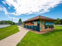 A small building with colorful graffiti art reading "Rainbow Park" stands beside a paved path on Rainbow Avenue, surrounded by green grass and fenced sports courts under a partly cloudy sky. - Beise Real Estate