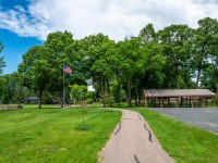 A paved pathway on Rainbow Avenue leads to a covered picnic shelter and a playground with an American flag, surrounded by green grass and trees under a partly cloudy sky in Maple Plain. - Beise Real Estate