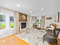 Bright living room with large windows on Old Crystal Bay Road in Orono, featuring a brick fireplace, armchairs, wicker chairs, decorative mirrors, and a patterned rug on a hardwood floor at 405 S. - Beise Real Estate