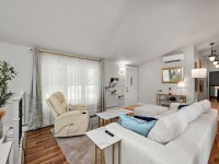Bright living room on Rainbow Avenue with a white sofa, recliner, coffee tables, wood flooring, wall-mounted shelves, large window with curtains, and a door leading to the hallway in Maple Plain. - Beise Real Estate