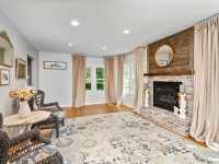 Bright living room at 405 S Old Crystal Bay Road features wicker chairs, a round table, large windows with beige curtains, a brick fireplace with wood mantel, and a decorative round mirror above the mantel. - Beise Real Estate